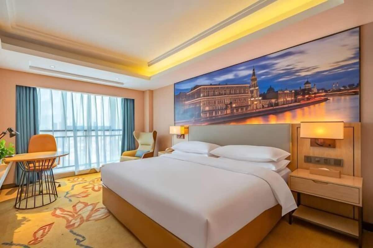 A hotel room with a large bed, a desk with a chair, a nightstand, and a painting of a cityscape.