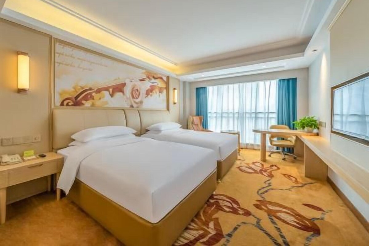 A hotel room with two beds, a desk, a chair, a window with curtains, and a painting on the wall.