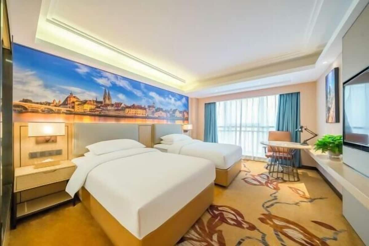 A hotel room with two beds, a desk, a chair, a large window with curtains, and a cityscape painting on the wall.