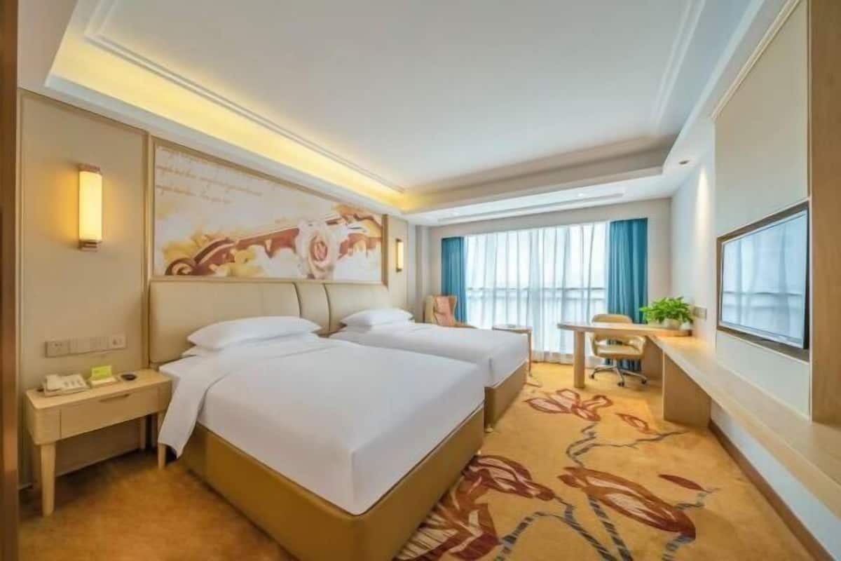 A hotel room with two beds, a desk, a chair, a small table, a plant, and a large window with curtains.