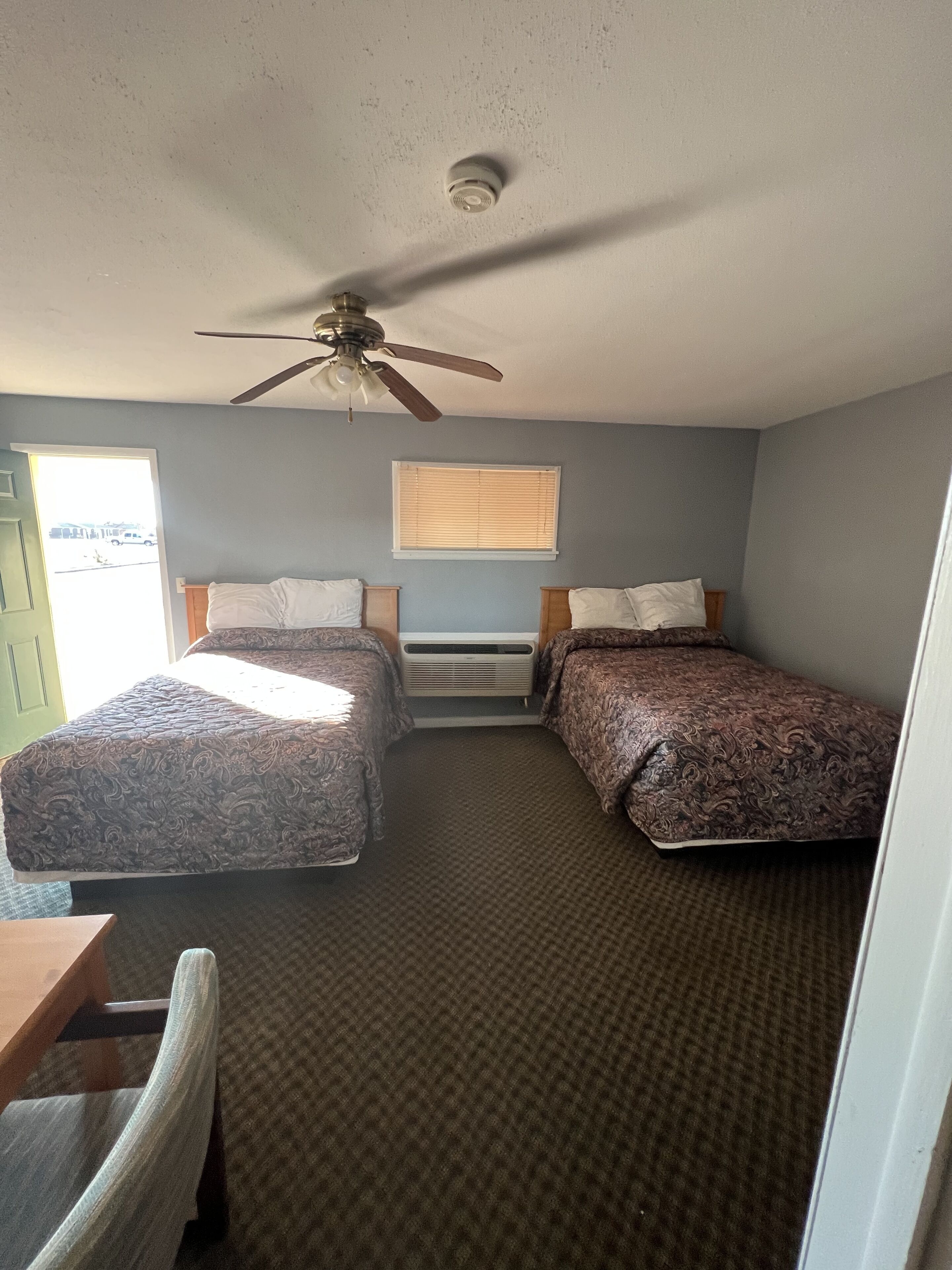 A room with two beds, a ceiling fan, a window with blinds, and a door.