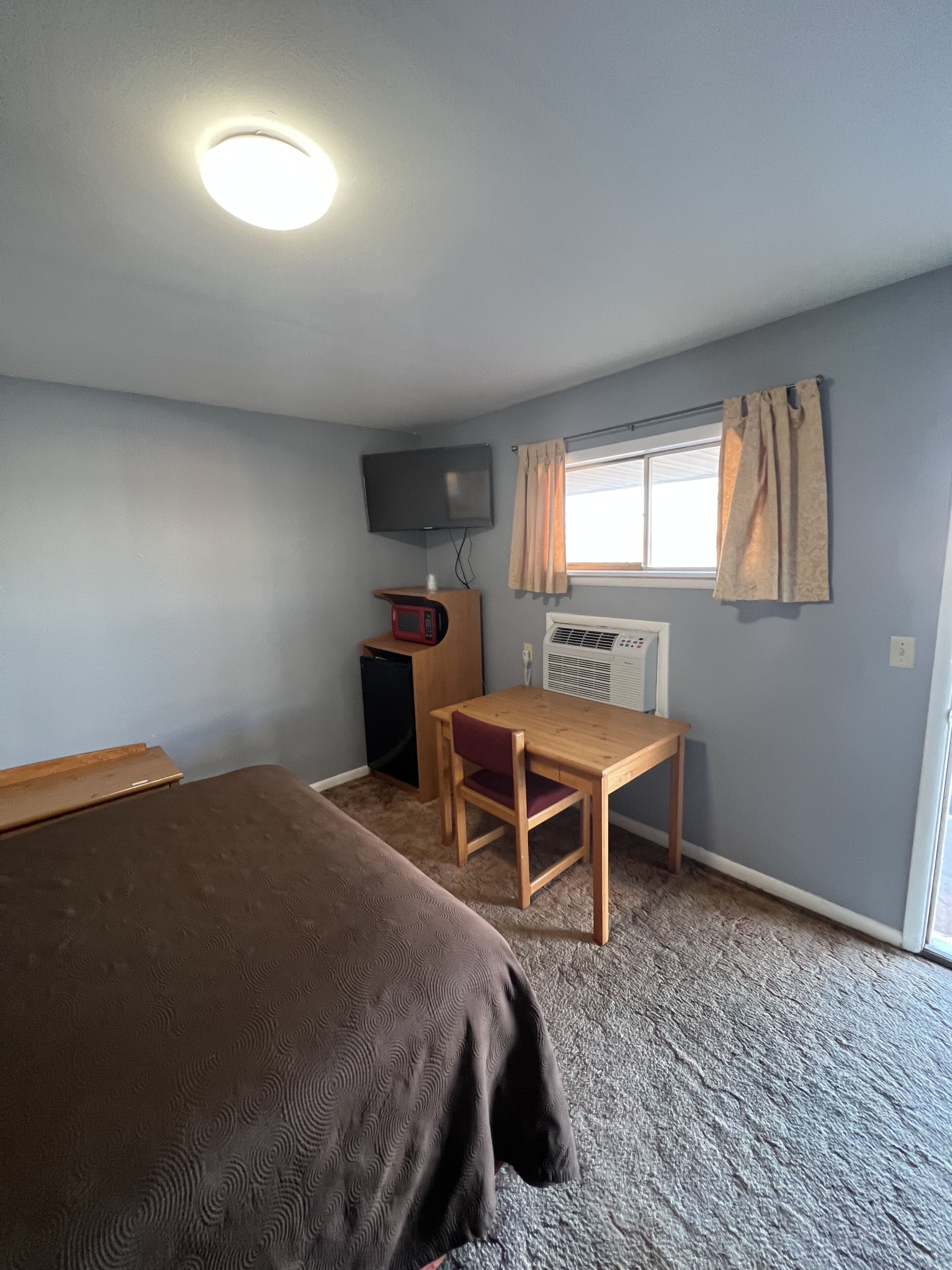Ein kleines Motelzimmer für eine Person mit Bett, Schreibtisch, Fernseher und einem Fenster mit Vorhängen.