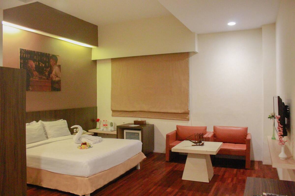 A hotel room with a bed, a small table, a sofa, a TV, and a window with a blind.
