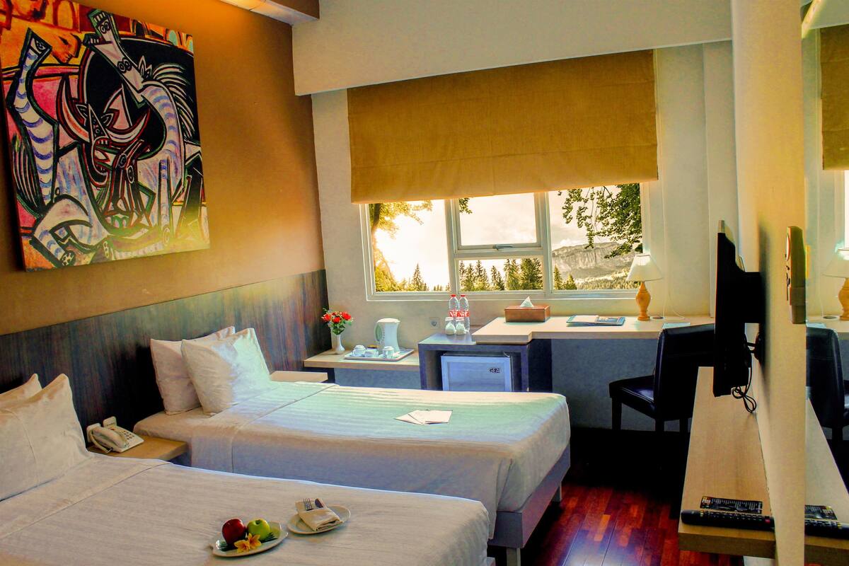 A hotel room with two beds, a desk, a chair, a TV, and a window with a view of trees.