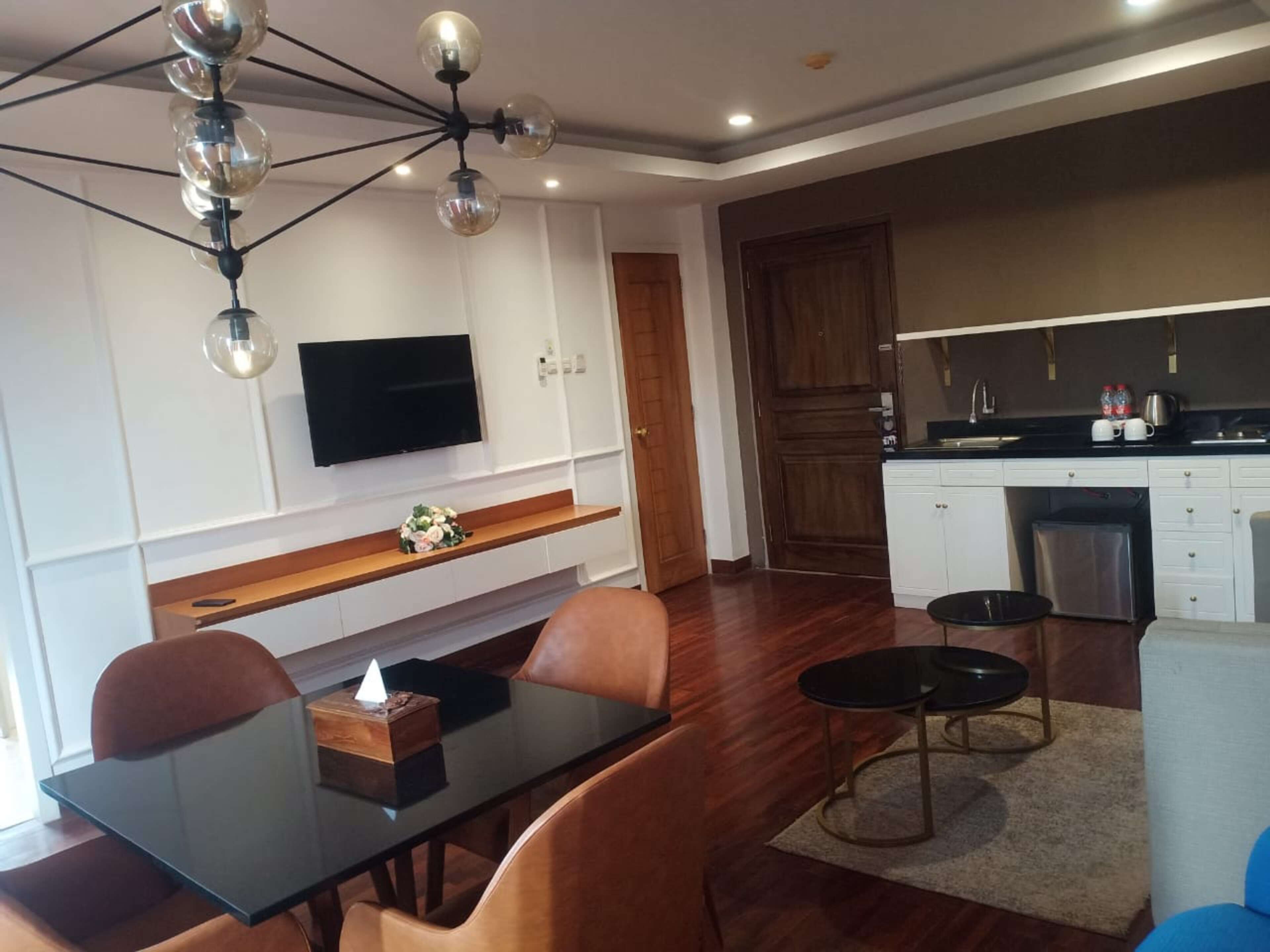 A modern living room with a glass dining table, leather chairs, a mounted TV, and a kitchen area with white cabinets and a wooden door.