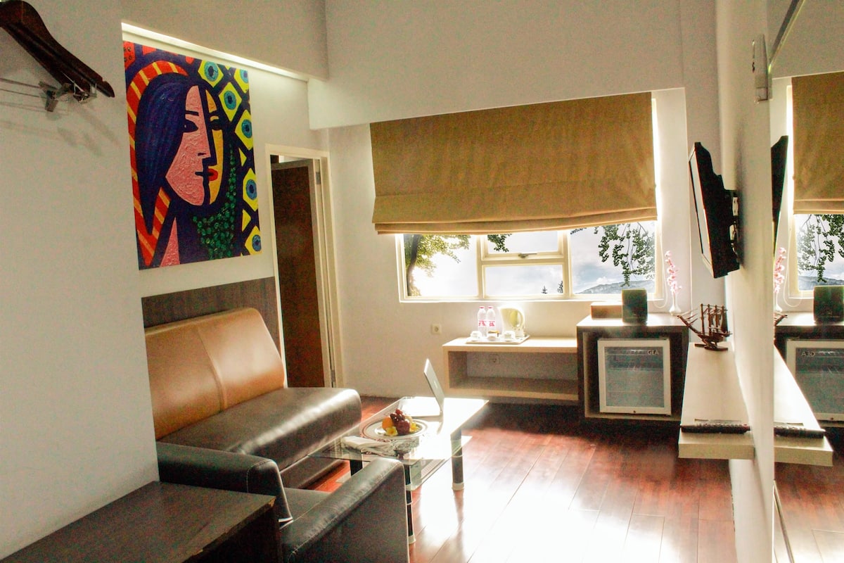 A living room with a sofa, a coffee table, a laptop, and a television. There is a window with a view of the outside.