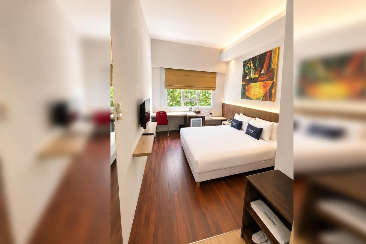 A modern hotel room with a large bed, a desk, a TV, and a window with a view of greenery.