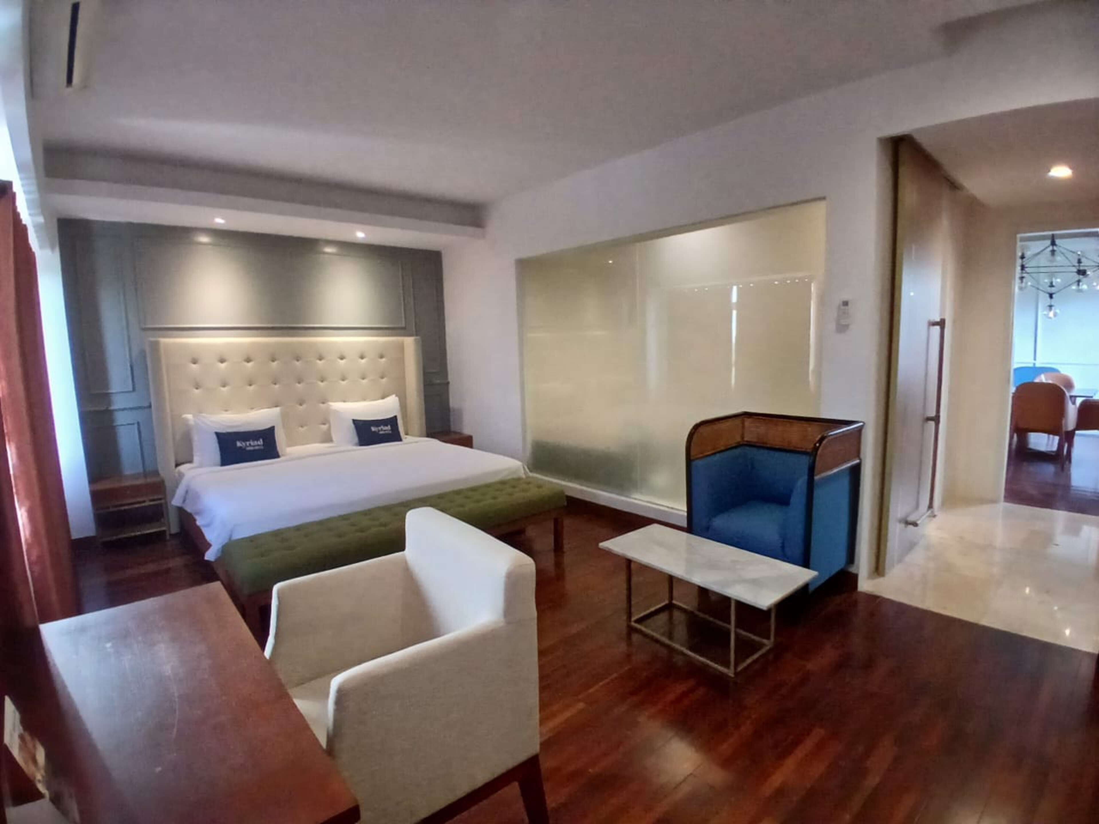 A hotel room with a large bed, a seating area with a table, and a view of the outside through a glass wall.