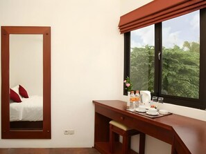 A hotel room with a bed, a desk with a mirror, a window with a view of trees, and a window blind.