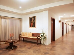 Un hall de hotel con un banco de madera, un jarrón con flores, un cuadro en la pared y un pasillo con puertas.