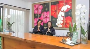 A reception desk with two individuals standing behind it, a large floral artwork on the wall, and a potted orchid on the counter.