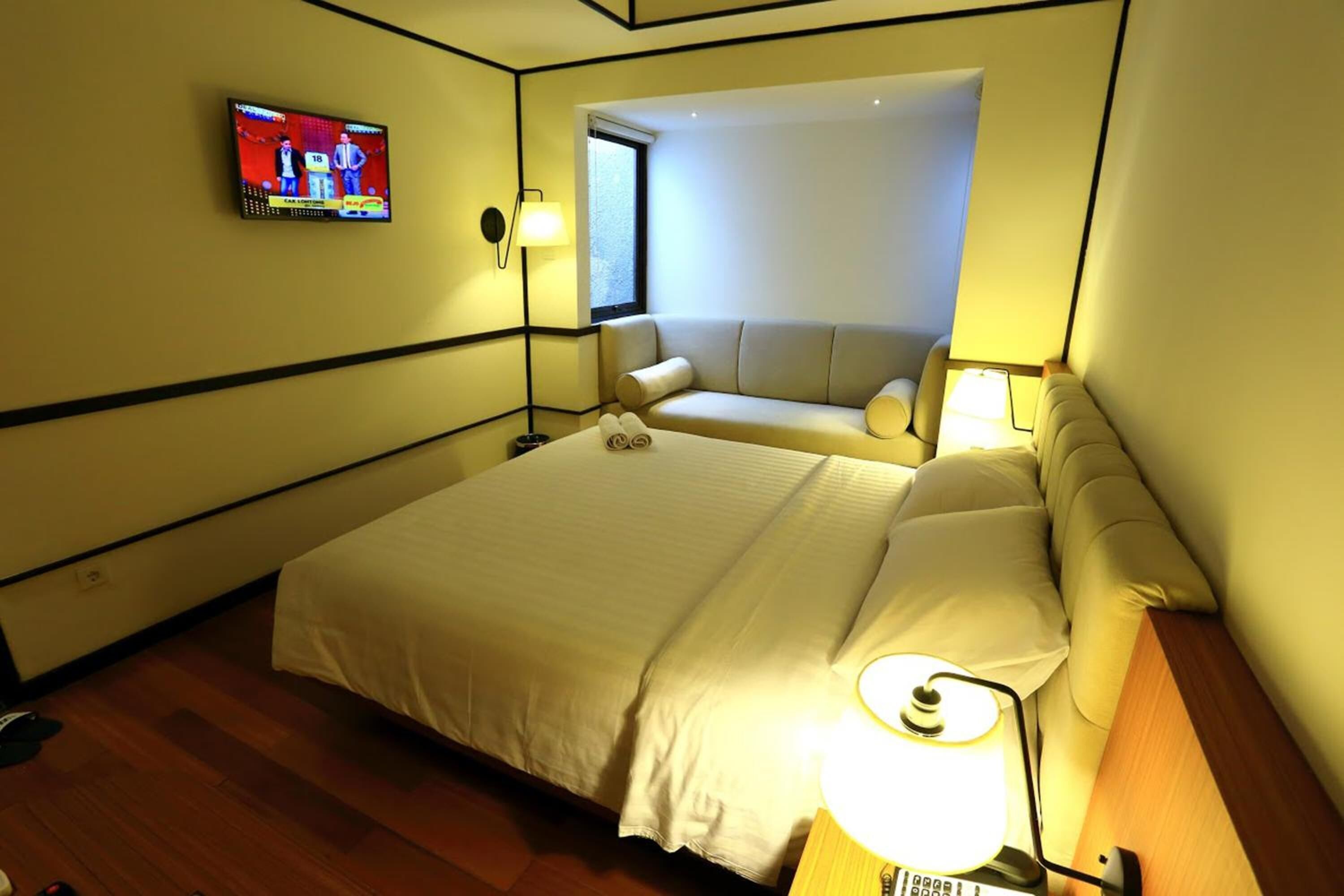 A hotel room with a bed, a TV mounted on the wall, a window, and a sofa.