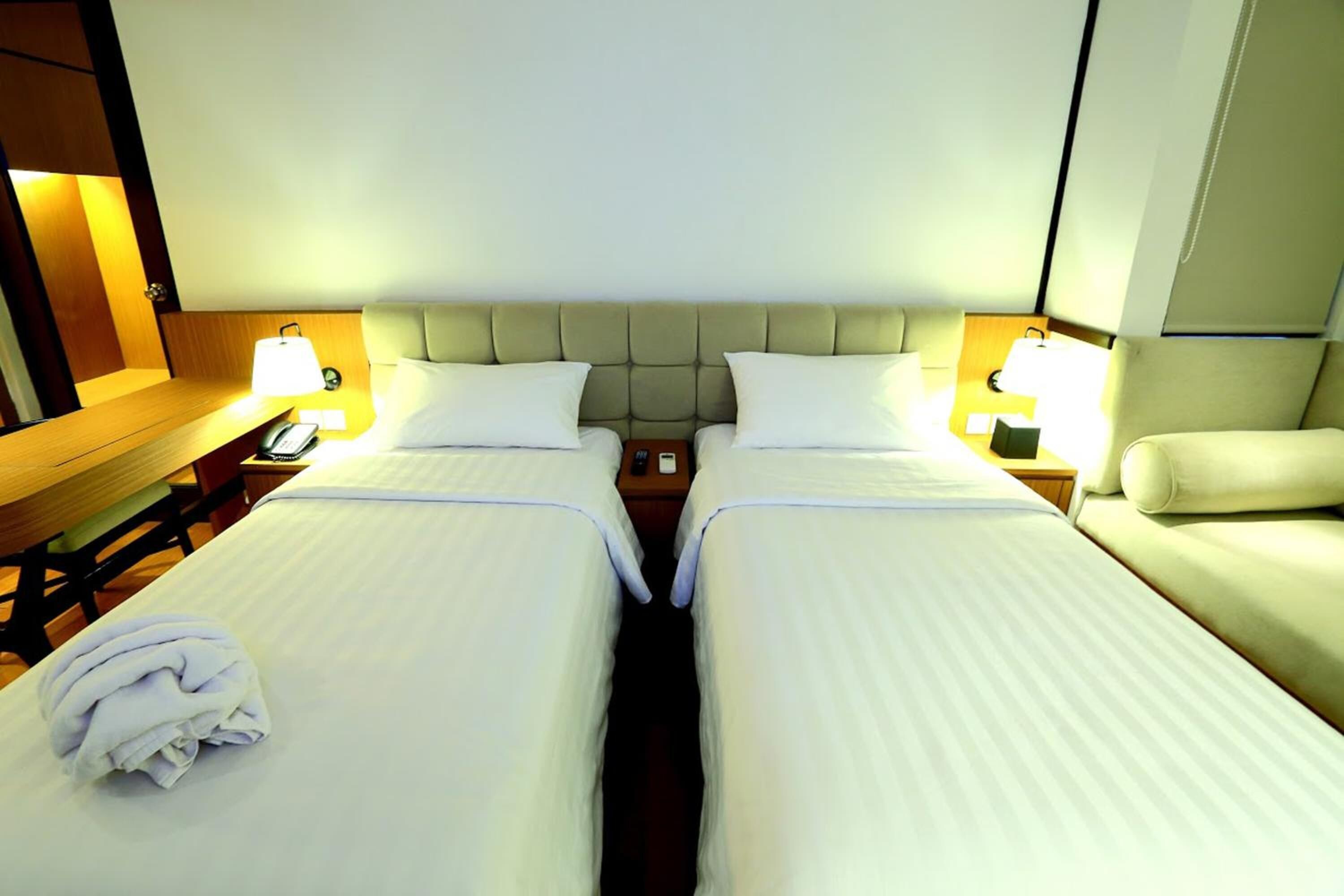 Ein Hotelzimmer mit zwei Betten, einem hölzernen Nachttisch, einer Lampe und einem gefalteten Handtuch auf dem Bett links.