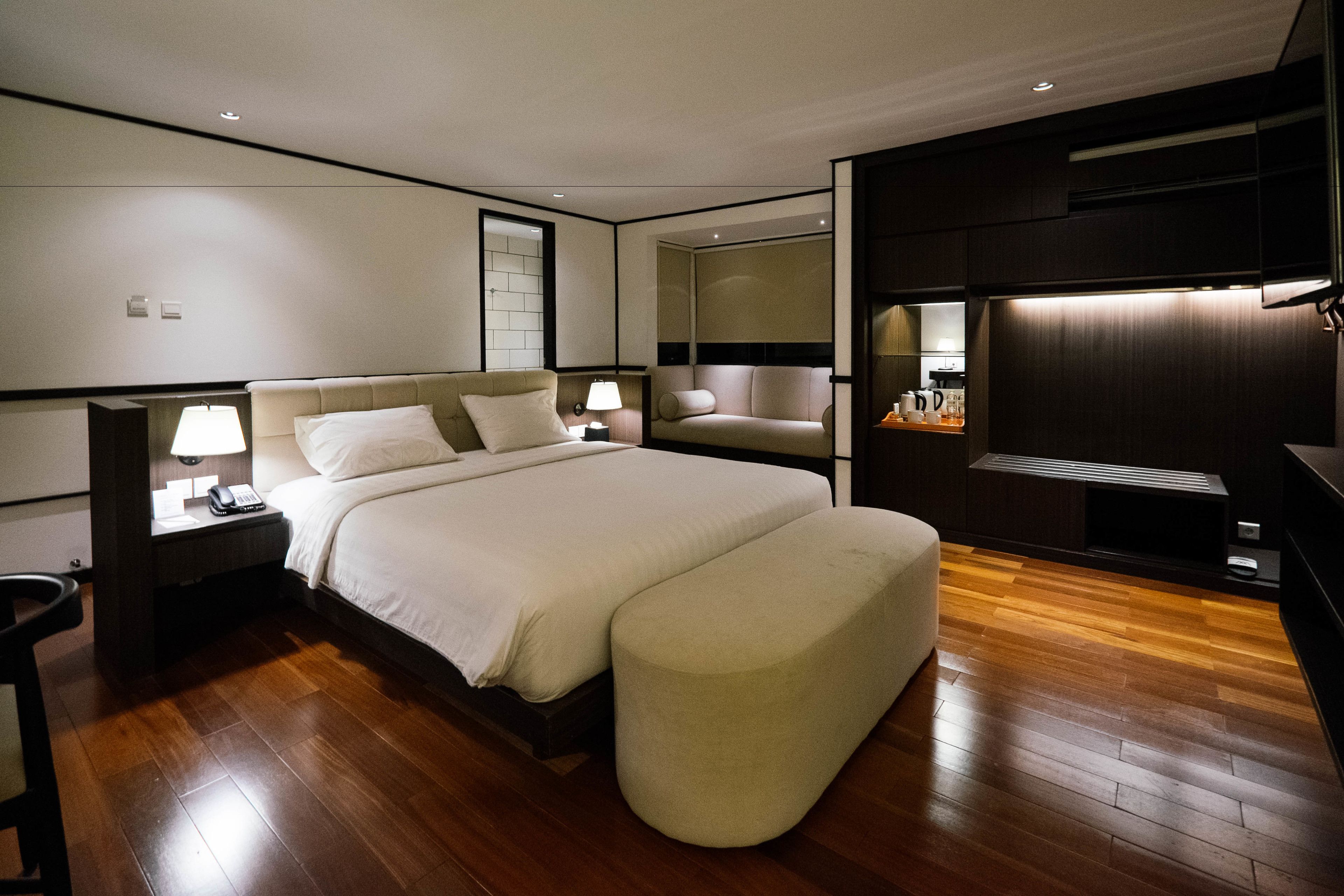 Una habitación de hotel moderna con una cama grande, una mesita de noche con lámpara, una zona de estar con un banco acolchado y un televisor integrado.