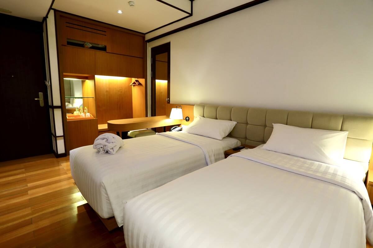 A hotel room with two beds, a wooden floor, a desk with a lamp, and a mirror.