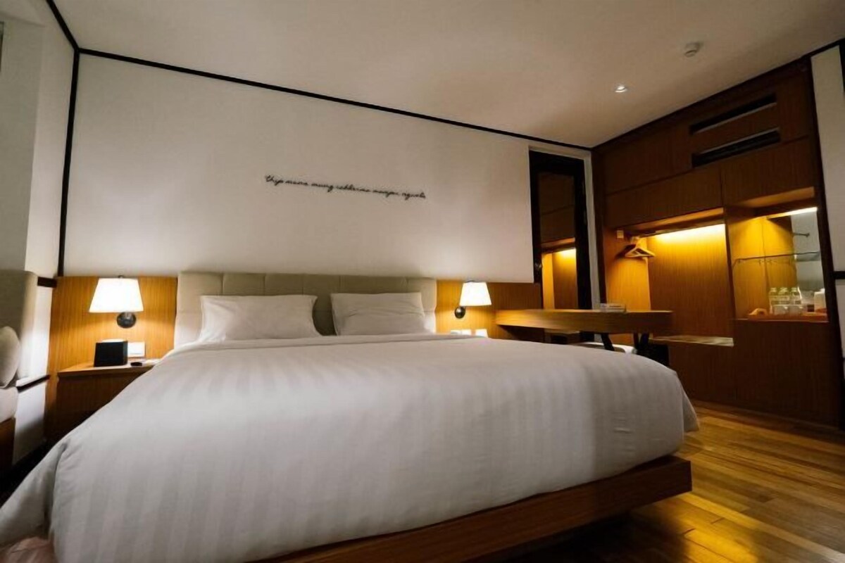 Moderni hotellihuone, jossa on suuri sänky, kaksi yöpöytää valaisimineen, puinen pääty ja seinälle kiinnitetty hylly, jossa on peili ja pyyheteline.