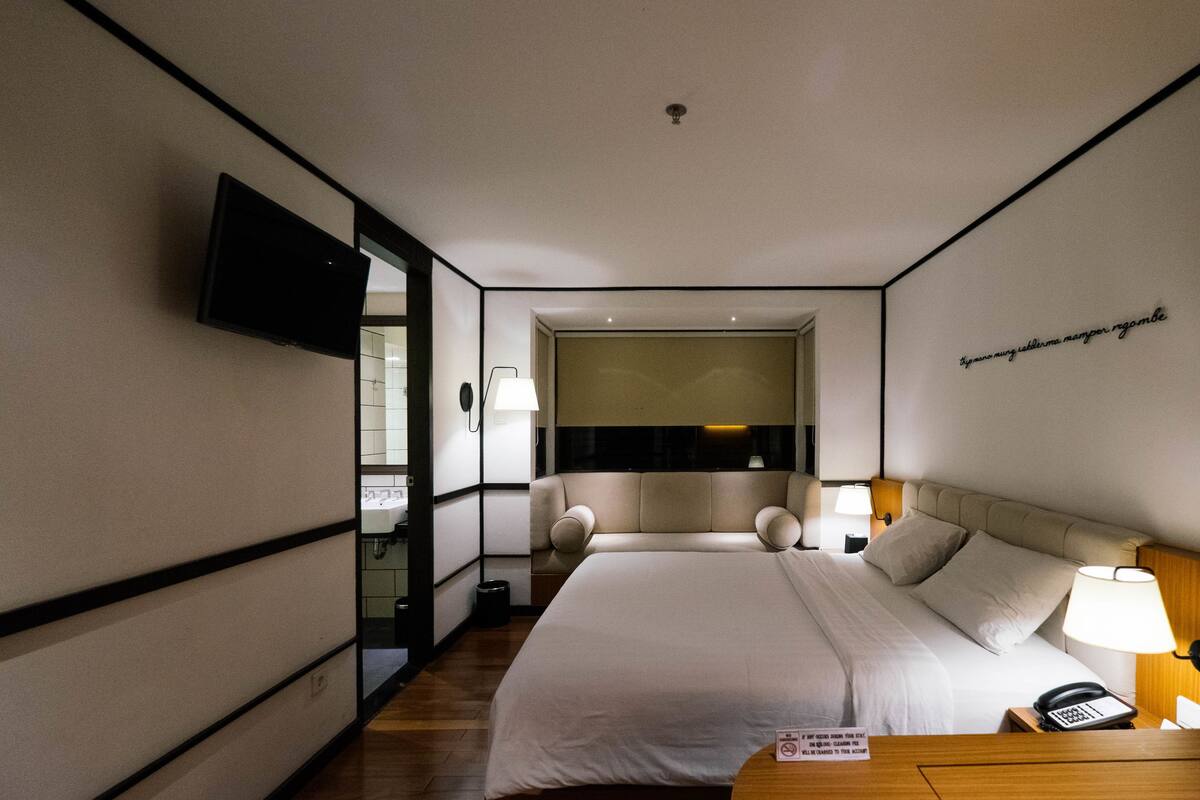 A modern hotel room with a large bed, bedside lamps, a flat-screen TV, and a view of the city through the window.