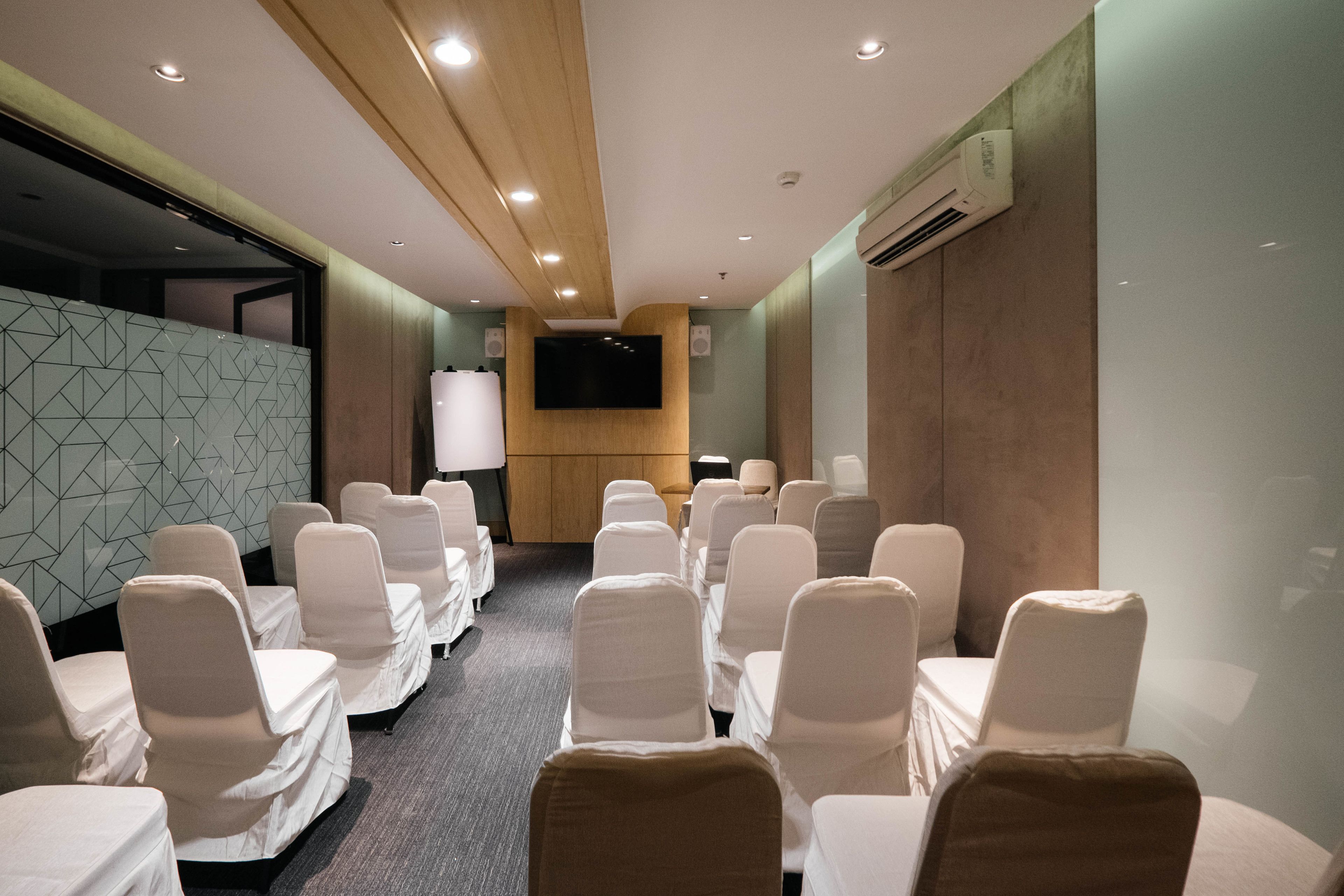 Uma sala de conferências com fileiras de cadeiras cobertas por toalhas brancas, uma TV de tela plana fixada na parede e um aparelho de ar condicionado.