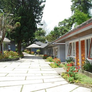 A row of single-story buildings with a central pathway, surrounded by greenery and trees. - Rudang Berastagi Hotel and Resort (Berastagi)
