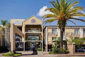 A hotel with a sign reading "PROTEA HOTEL," a two-story structure with a balcony, and palm trees in the foreground.