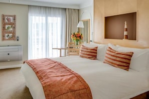 A hotel room with a large bed, a desk with a chair, a lamp, and a wall-mounted shelf with a decorative item.