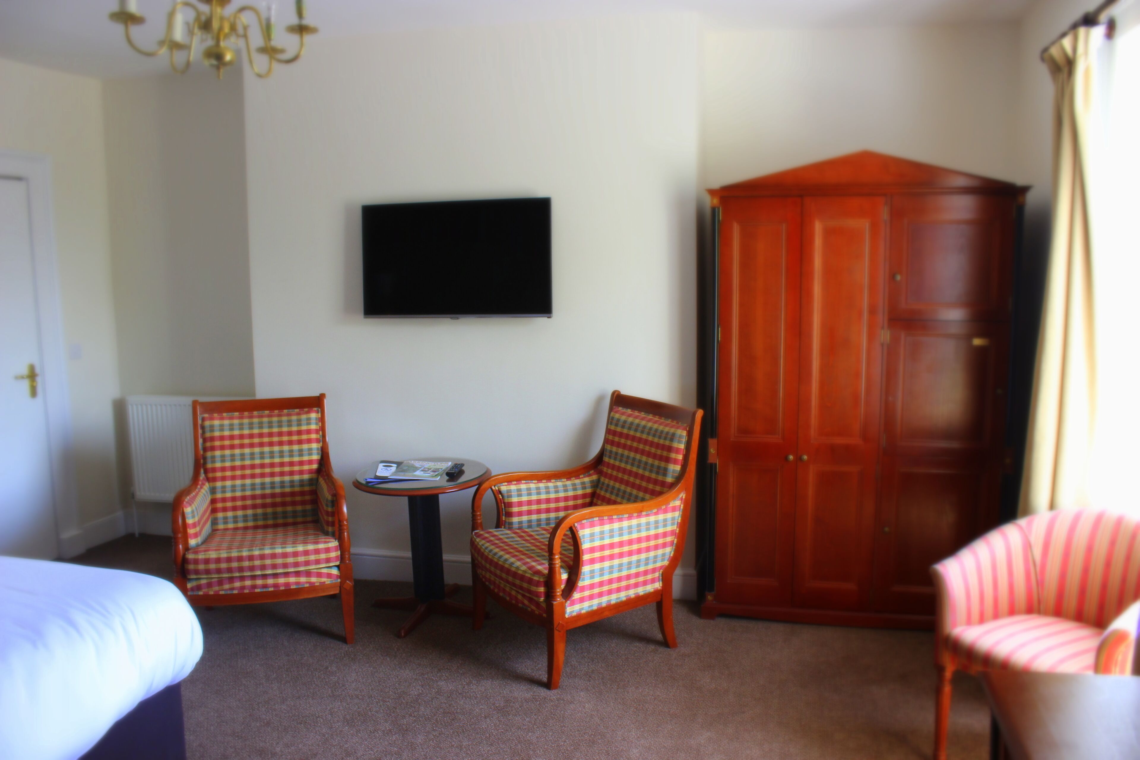 Habitación de hotel con dos sillones a cuadros, una mesita redonda, un televisor en la pared y un armario de madera.