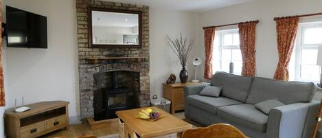 A living room with a stone fireplace, a grey sofa, wooden chairs, a dining table with a plate of food and a glass of orange juice, and a TV mounted on the wall.