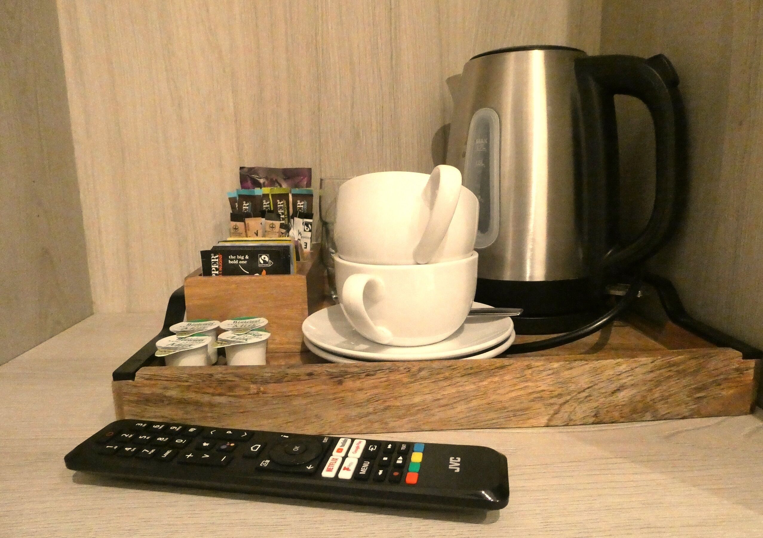 Un control remoto, una taza blanca con una cuchara, una cafetera y una bandeja con varios objetos sobre una superficie de madera.