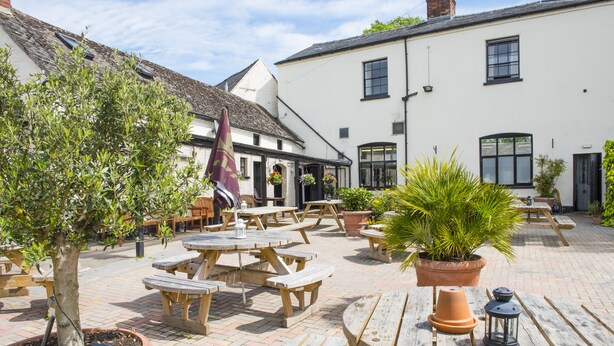 An outdoor patio area with wooden tables and benches, potted plants, and a white building with windows and doors in the background. - The Frocester (Stonehouse)