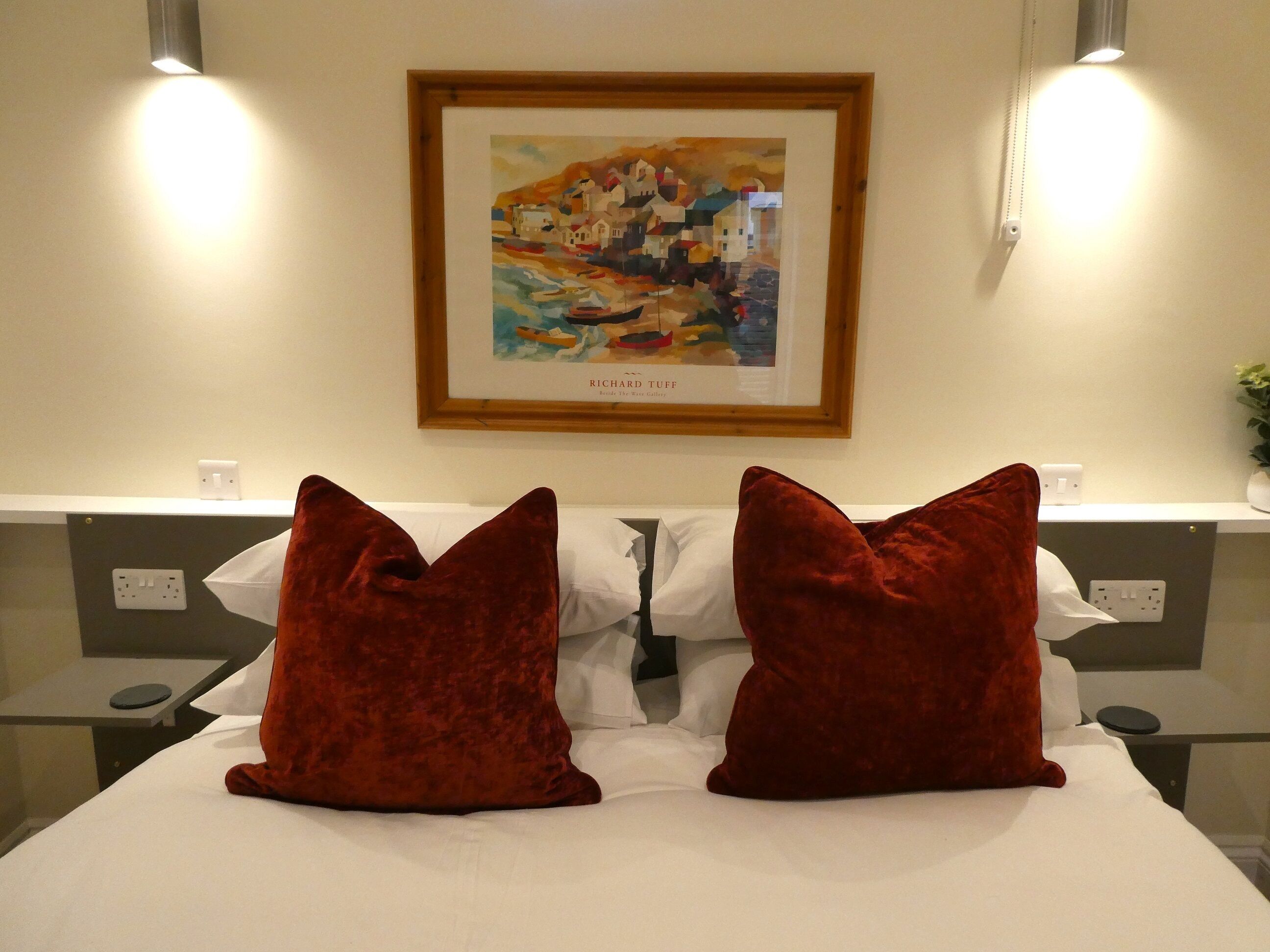 A hotel room with a bed, two red pillows, a framed artwork on the wall, and bedside tables with lamps.