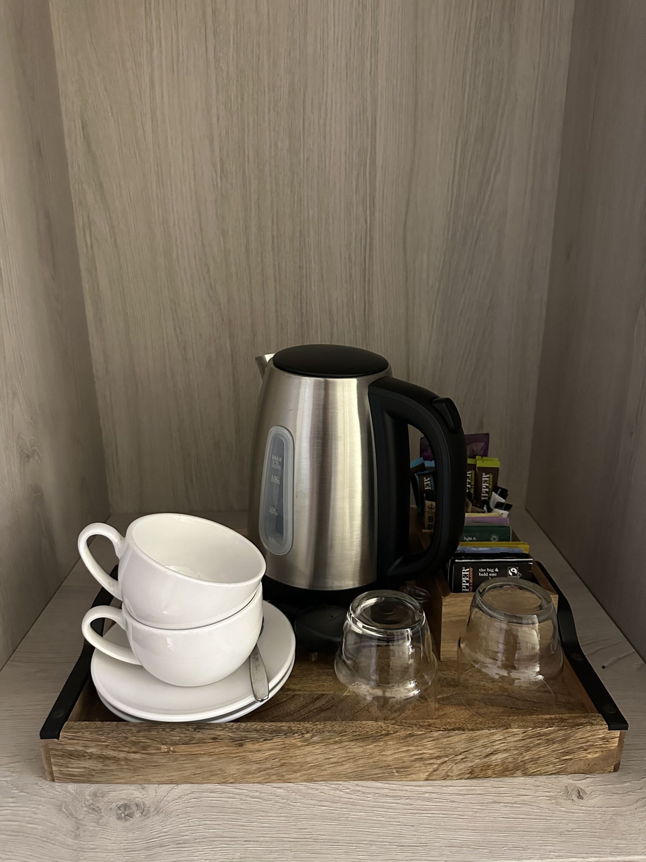 A stainless steel kettle, two white cups, and a glass on a wooden tray.