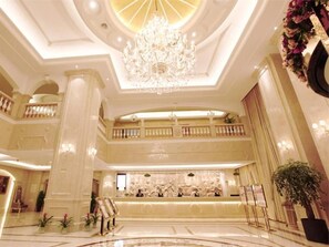 A grand lobby with a central reception desk, a chandelier, and a high ceiling.