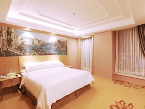 A hotel room with a large bed, a nightstand, a painting of a cityscape, a window with curtains, and a carpet with a floral pattern.