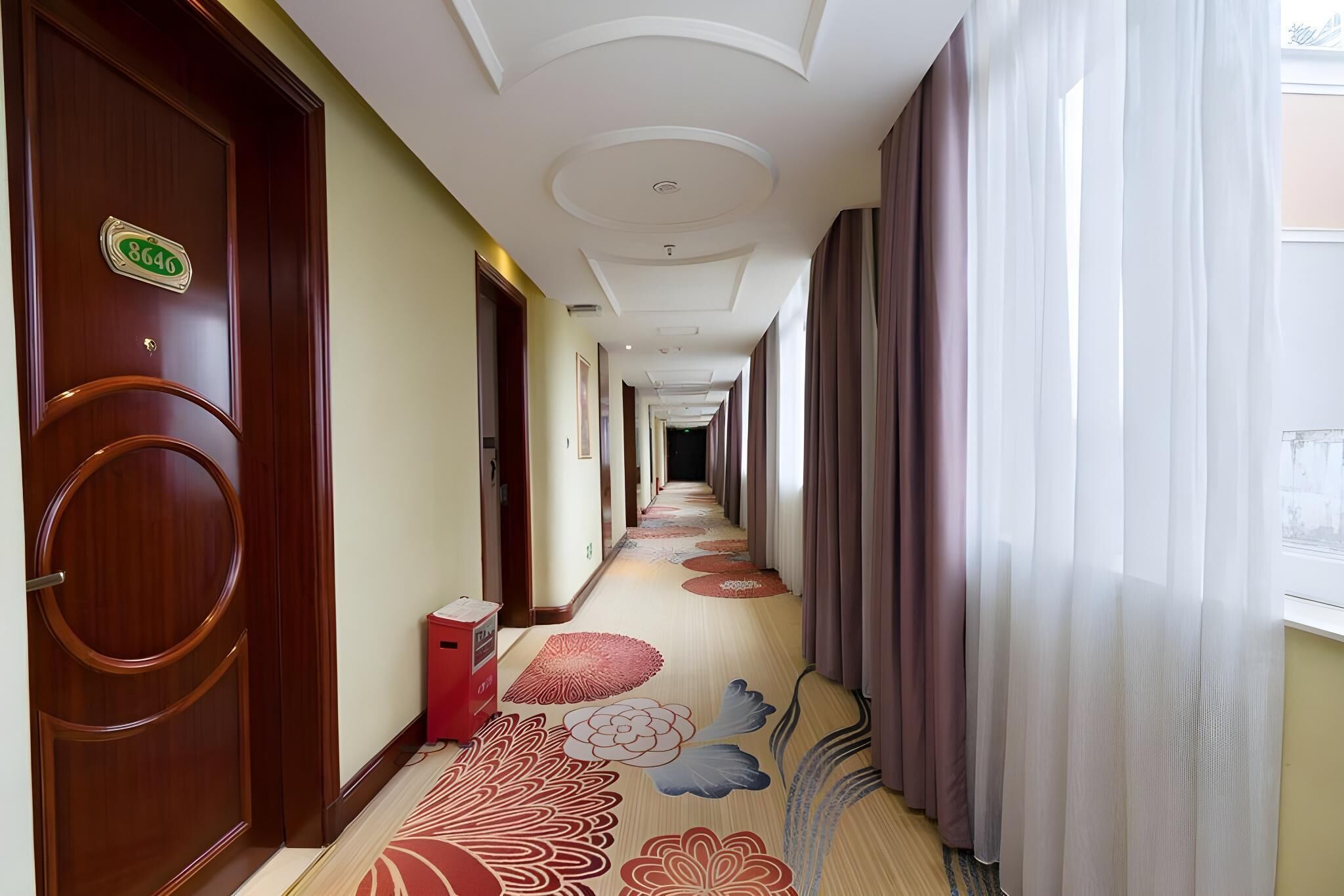 A hotel hallway with a wooden door, a red trash bin, and a carpet with floral patterns.