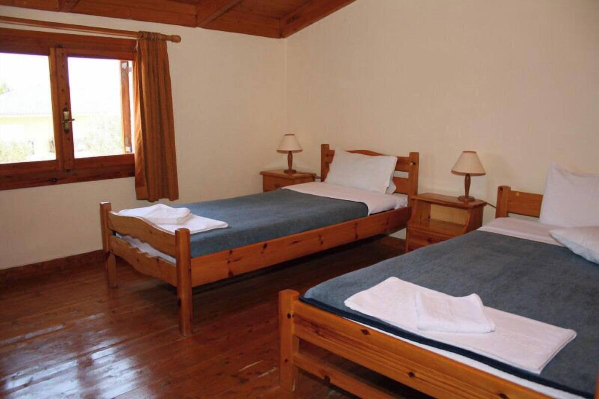 Una habitación con dos camas de madera, cada una con una mesita de noche y una lámpara. Se ve una ventana con cortinas a la izquierda.