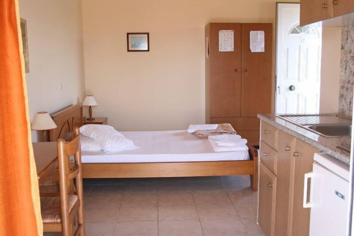 Una habitación pequeña y ordenada con una cama individual, un armario de madera, una mesita con una silla y un rincón con kitchenette.