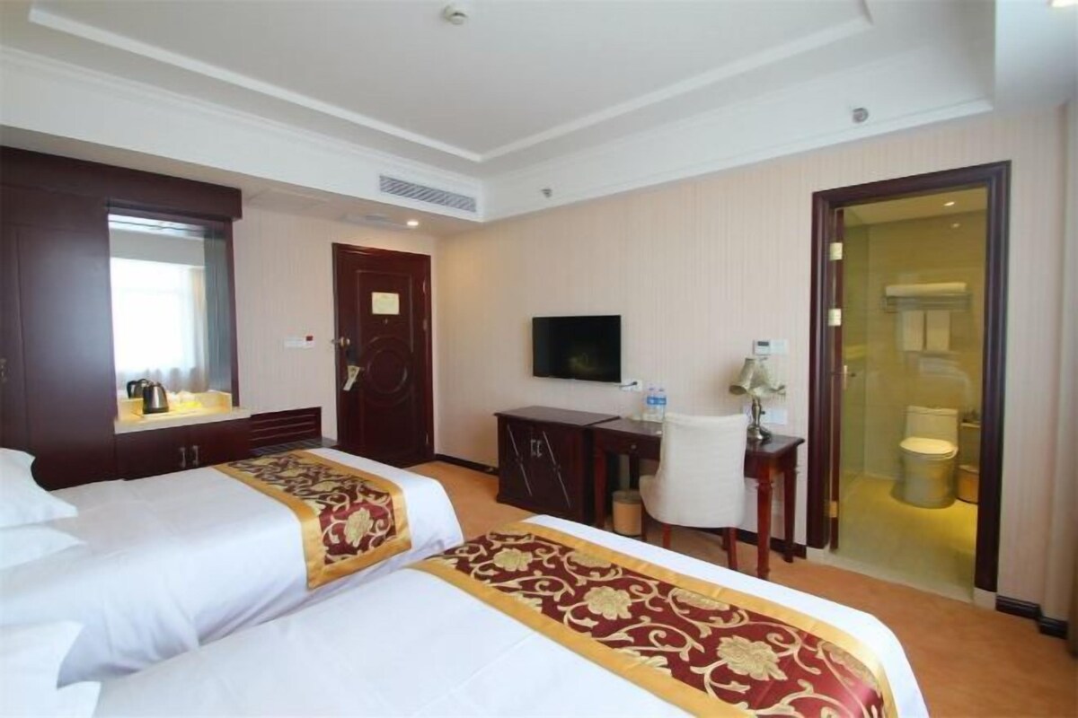 A hotel room with two beds, a desk, a television, and a bathroom visible through a glass door.