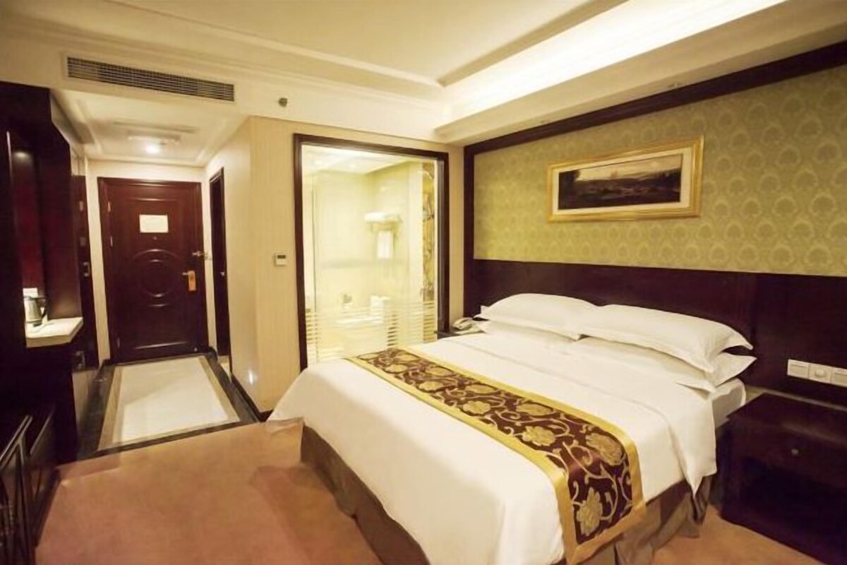 A hotel room with a large bed, a bedside table, a nightstand, a wall-mounted TV, and a door leading to a bathroom.