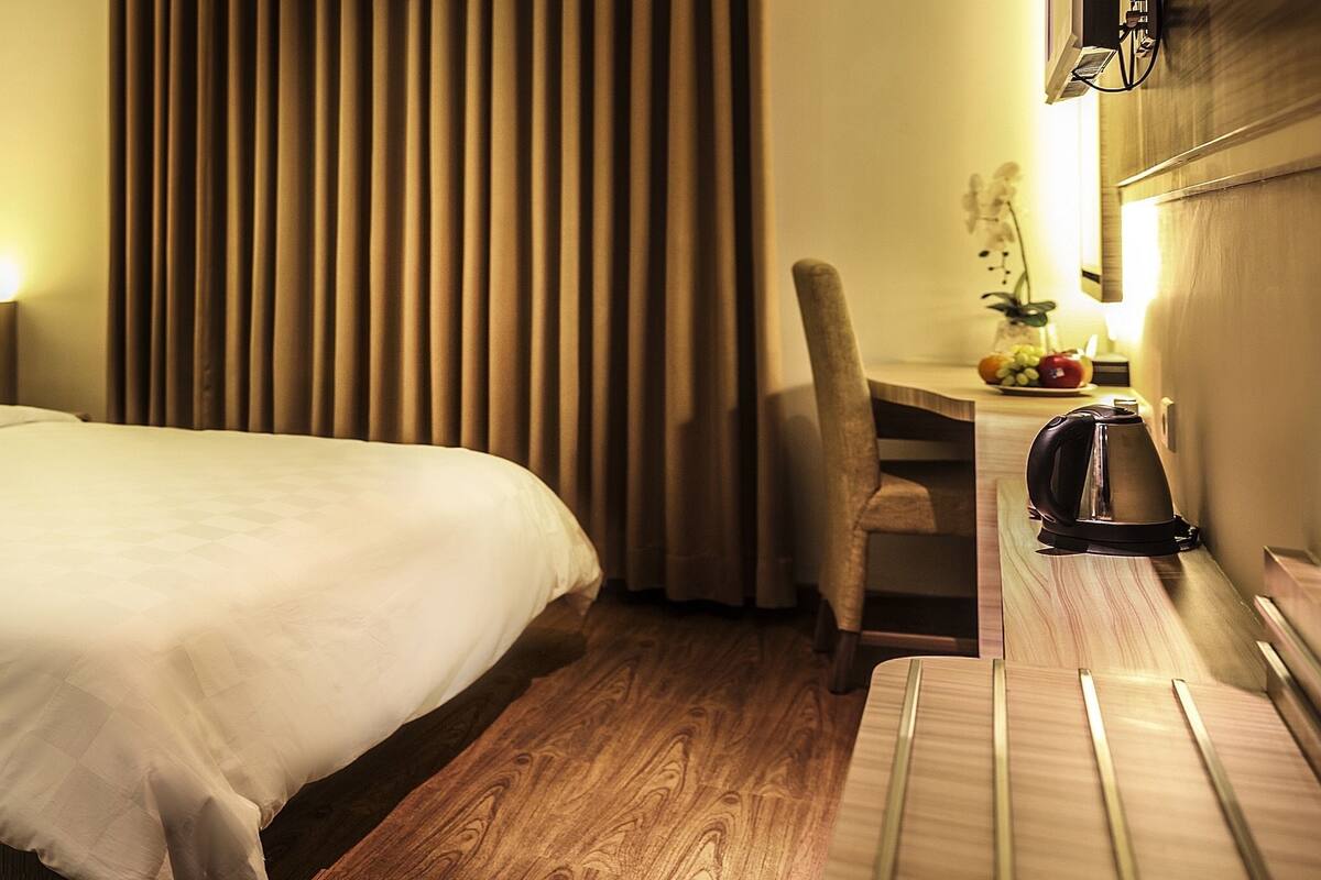 A hotel room with a bed, a desk with a chair, a coffee machine, and a vase with flowers.
