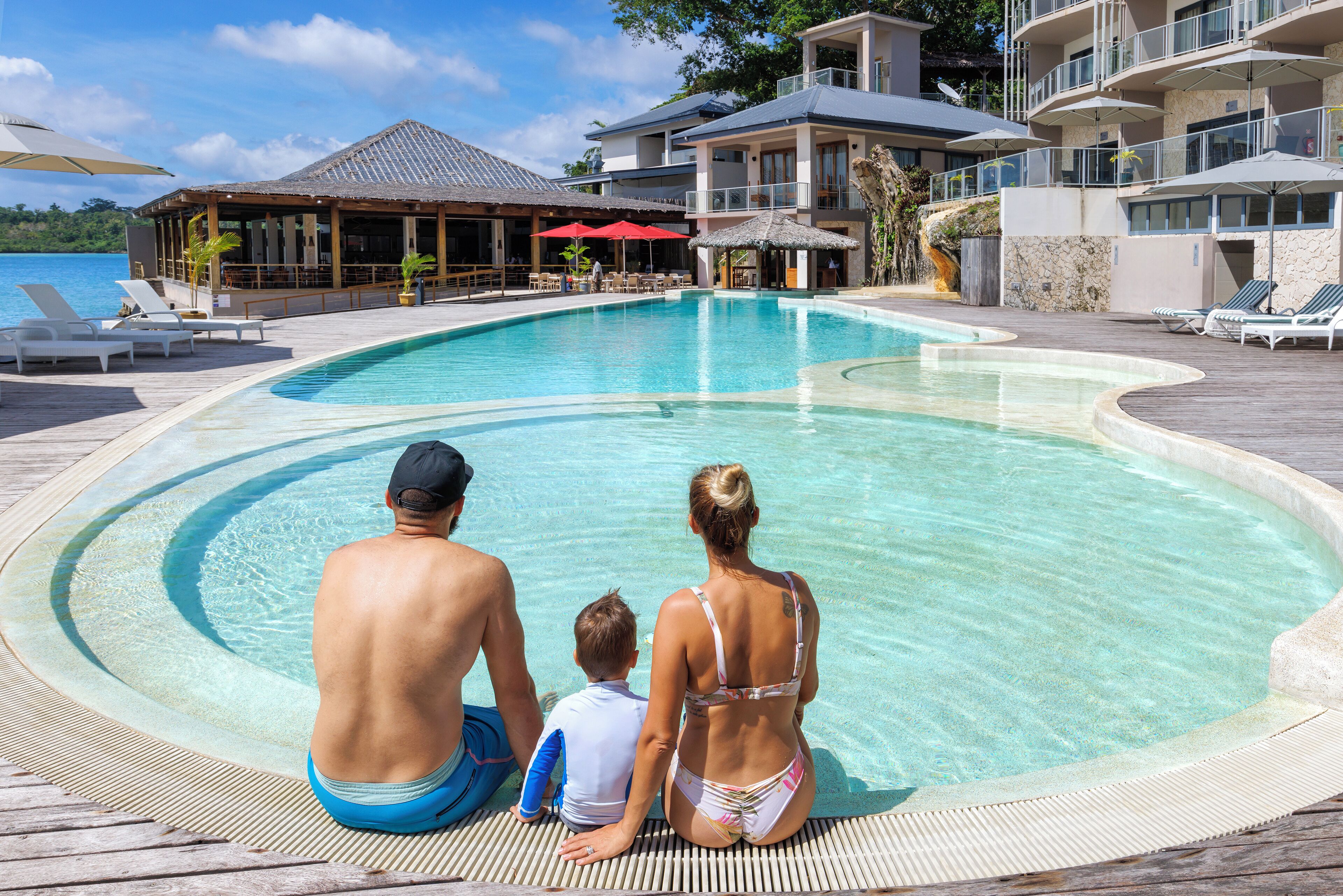 A family by a pool with a view of a resort building and the sea.