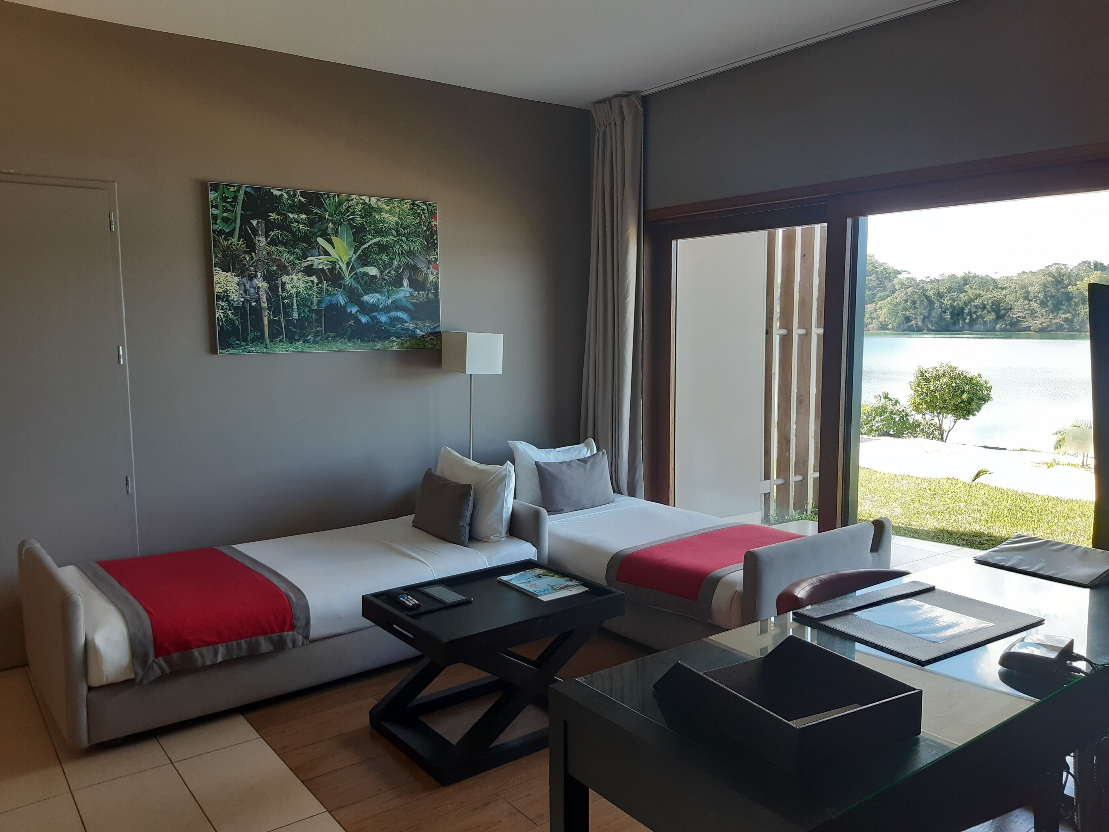 A modern hotel room with a large window, a sofa, a coffee table, and a painting of a tropical landscape.