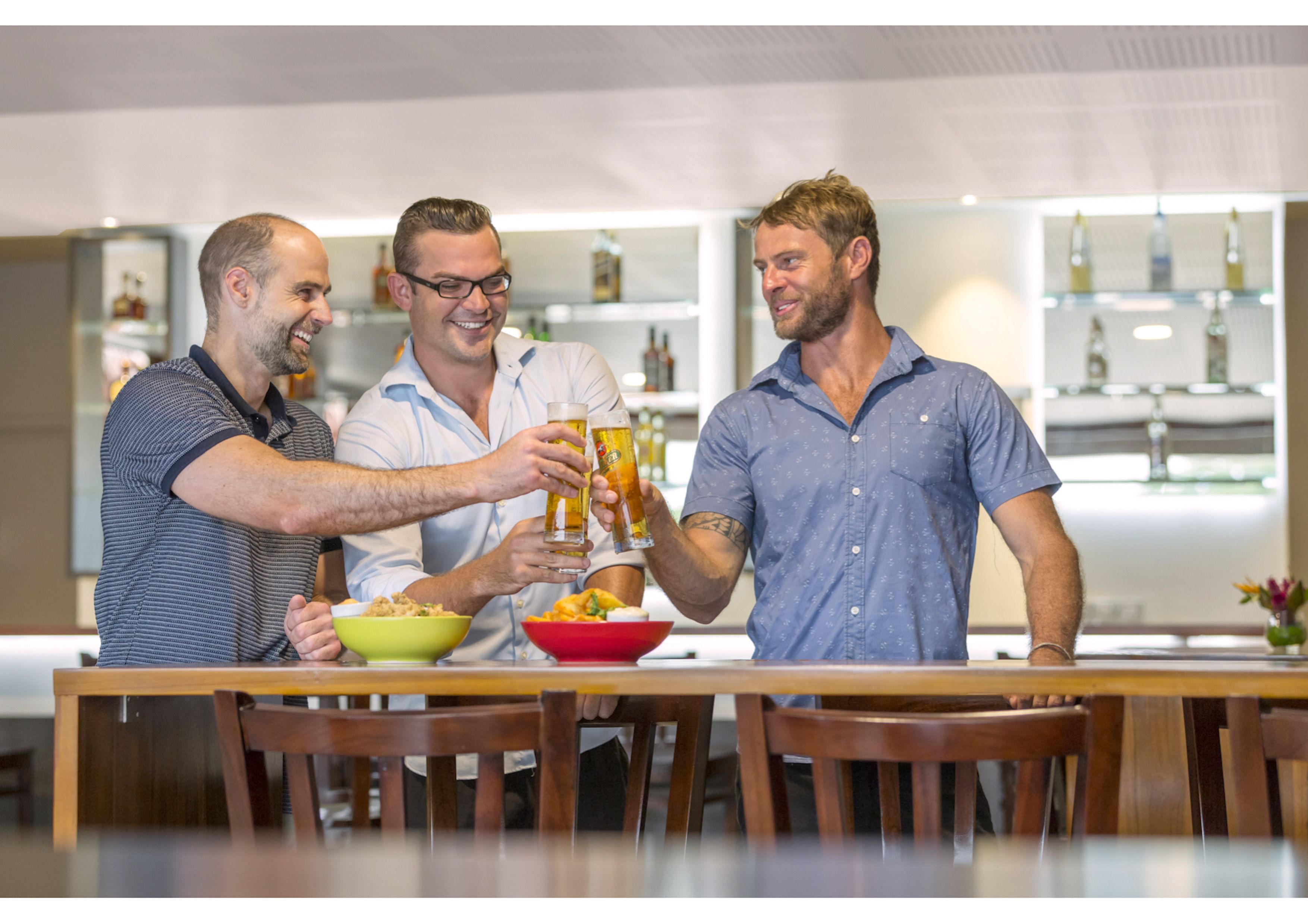 Un grupo de personas brindando con vasos de cerveza en una mesa de madera en un interior moderno con estanterías al fondo.