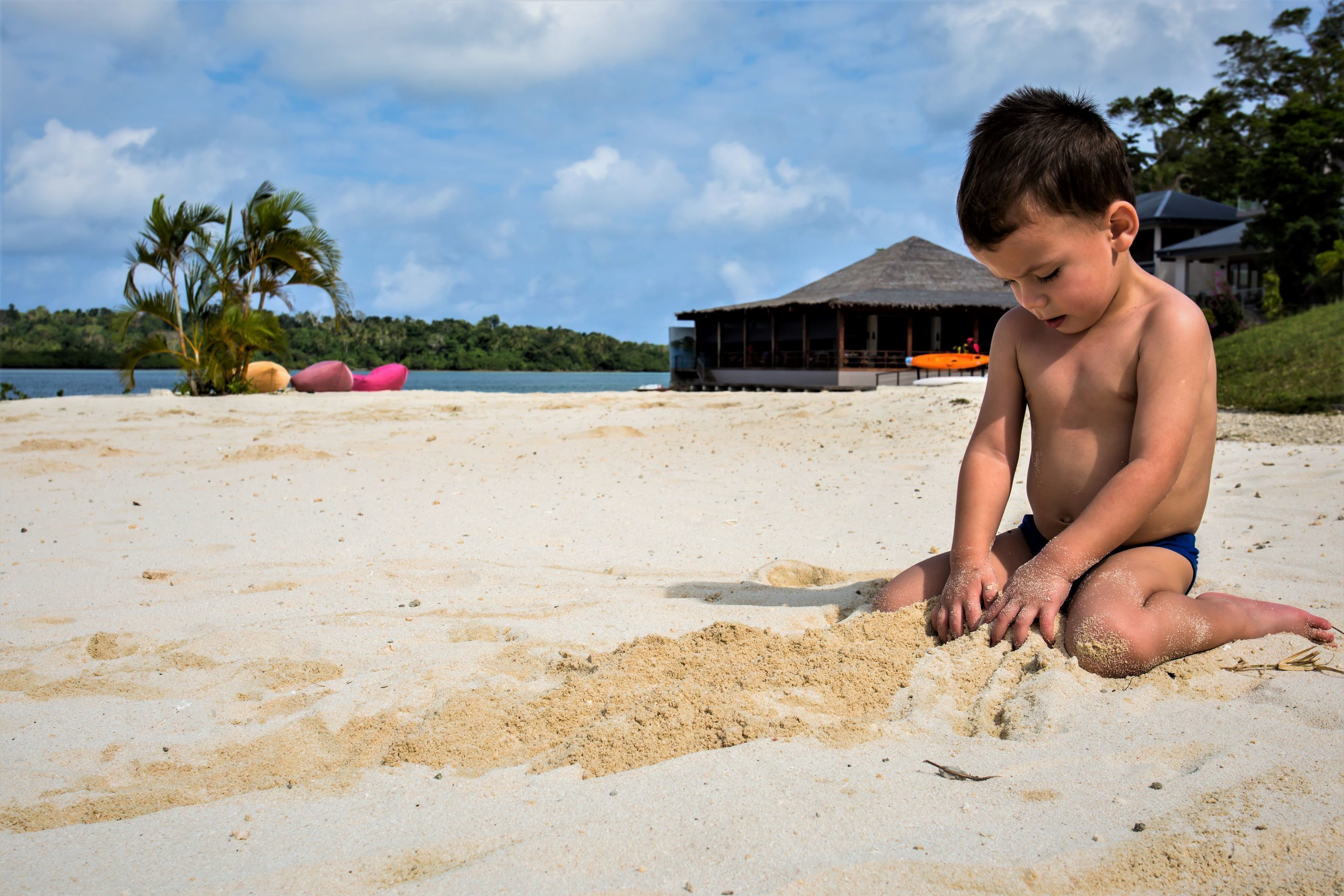 Escena de playa con un niño jugando en la arena, una estructura de techo de paja al fondo y palmeras.