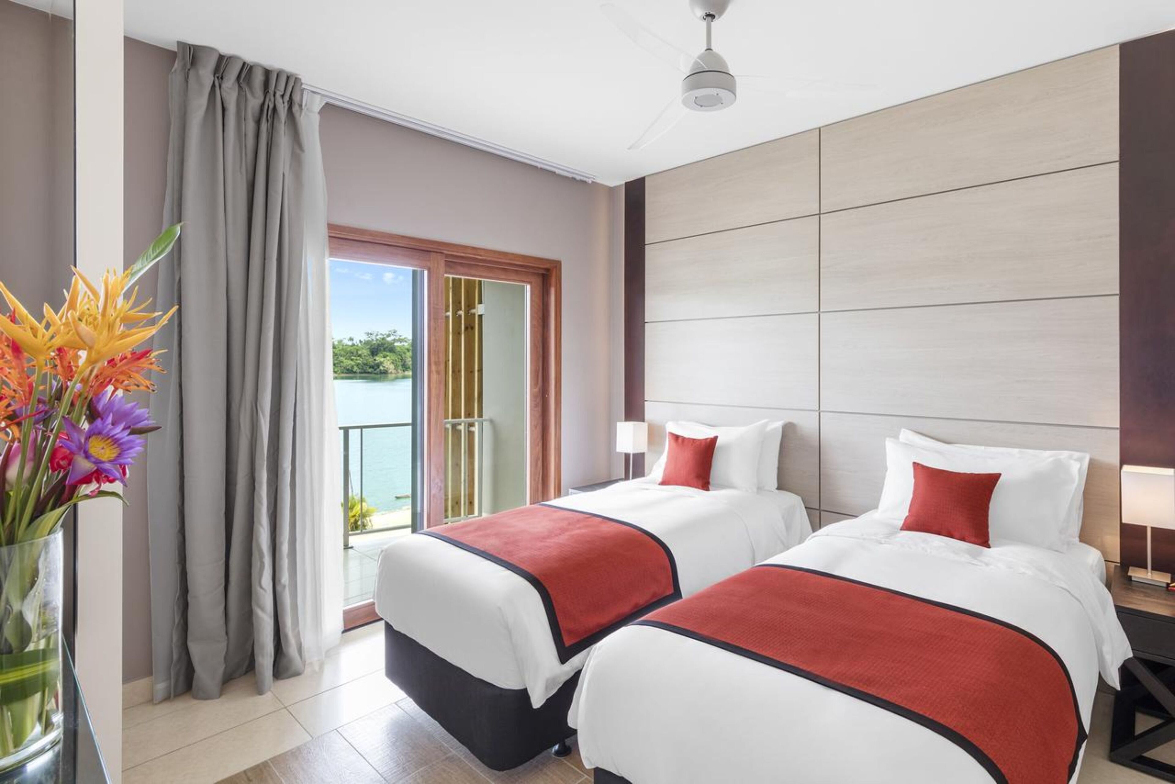 Une chambre d’hôtel avec deux lits, un balcon donnant sur la mer et un vase de fleurs placé à gauche.