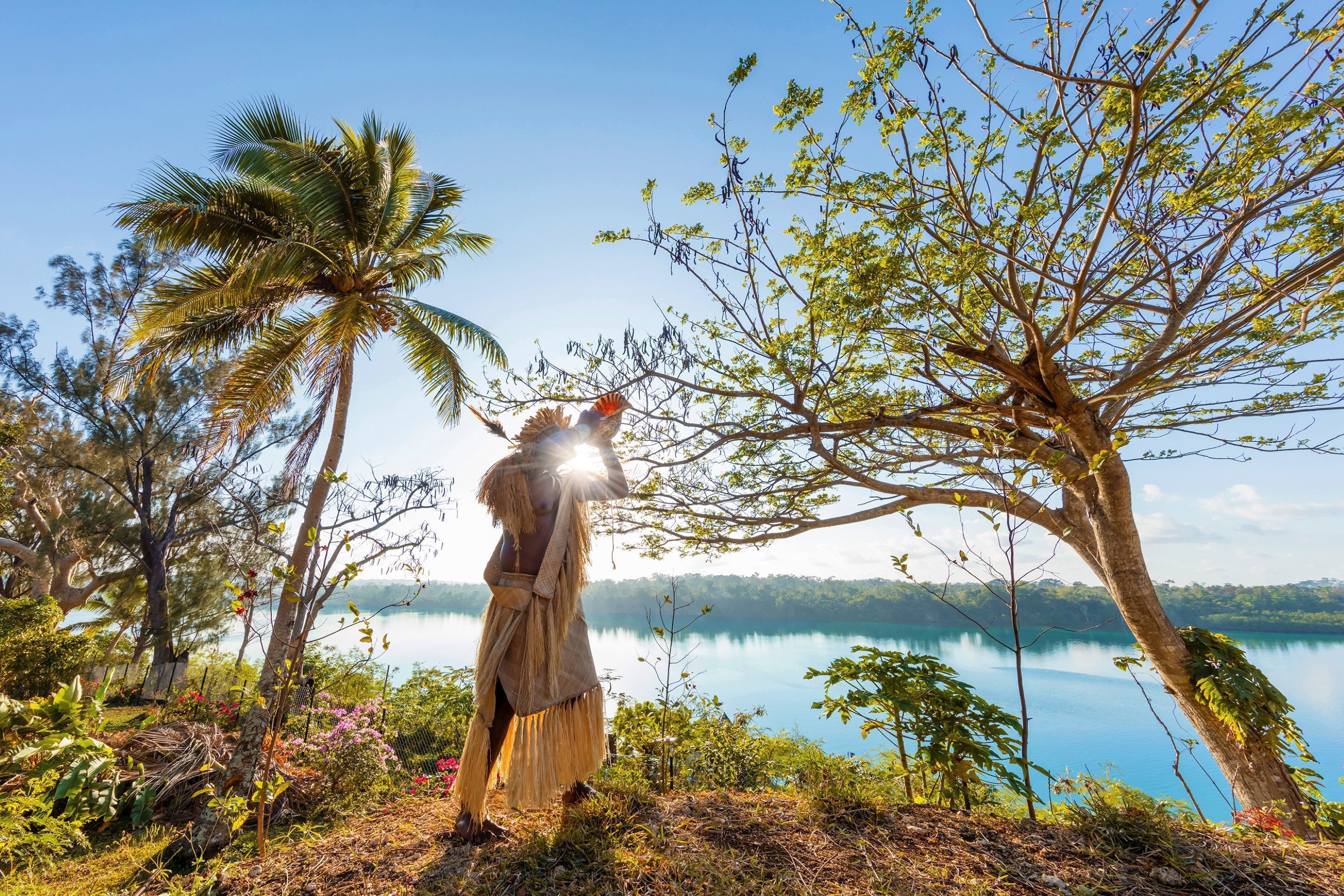 A person in traditional attire standing by a tree with a lake in the background.