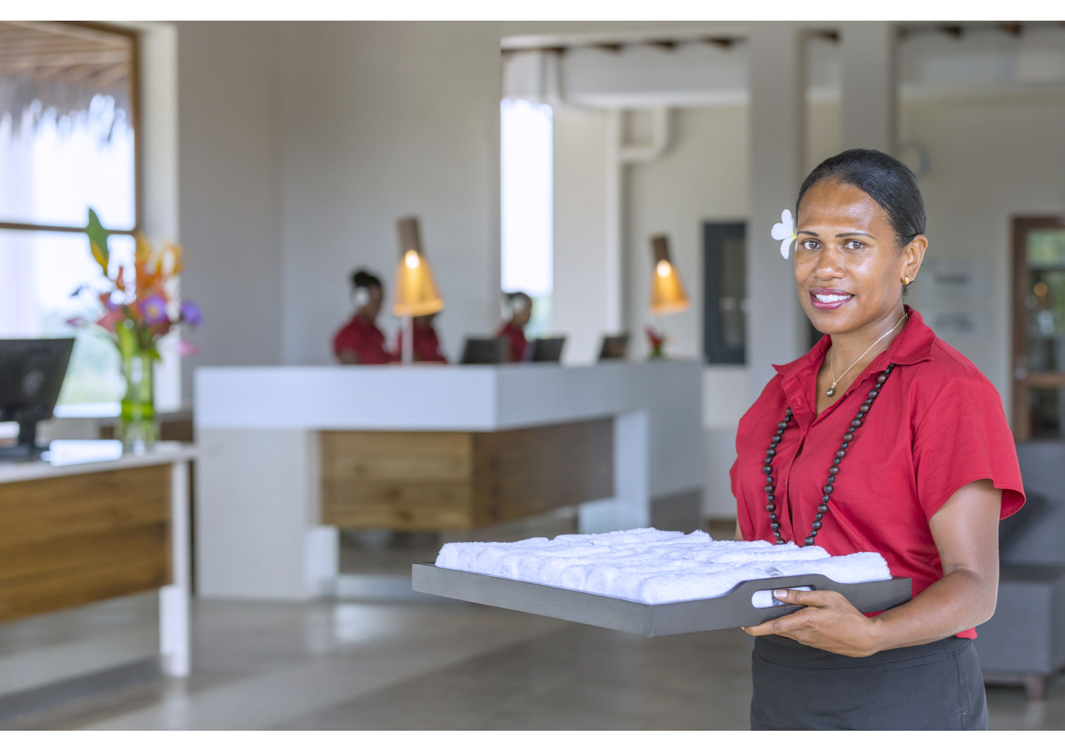 Zona de recepción de un hotel con diseño moderno, mostrador de recepción y un miembro del personal sosteniendo una bandeja con un objeto blanco.