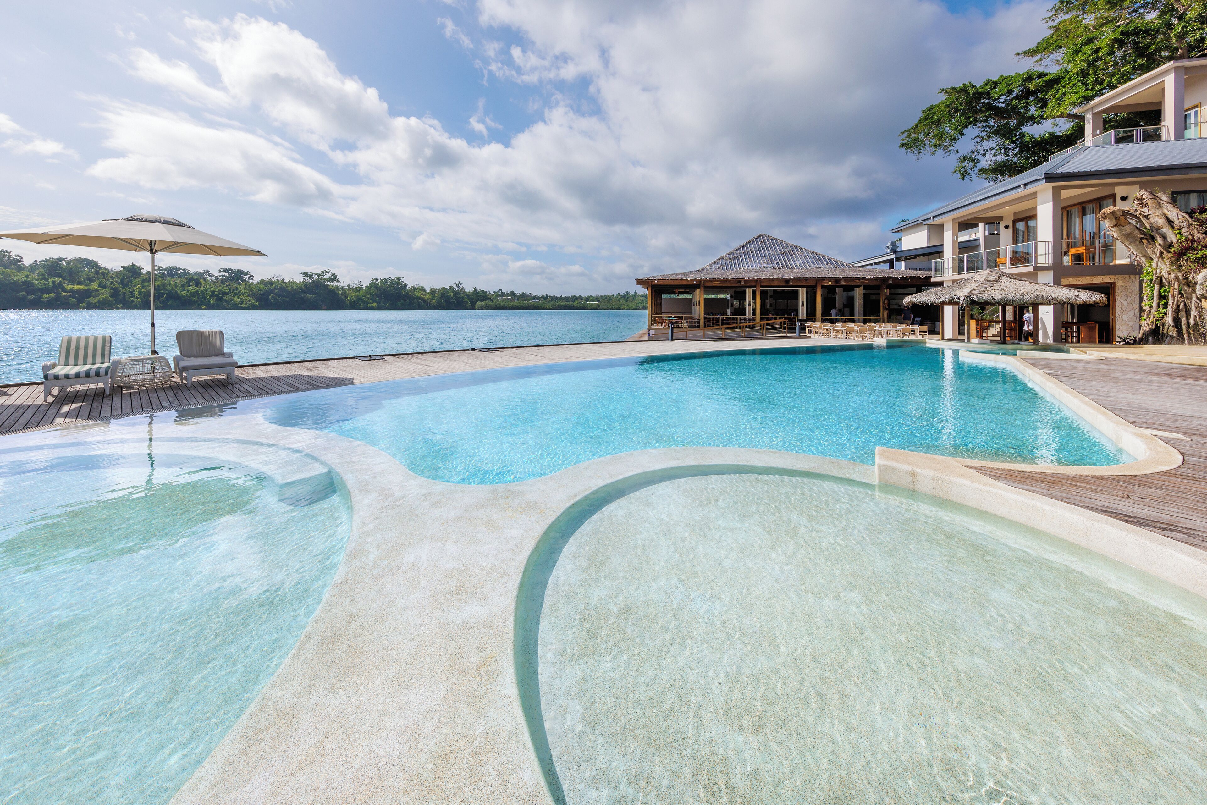 A clear swimming pool with lounge chairs and umbrellas, surrounded by a wooden deck and overlooking a body of water.