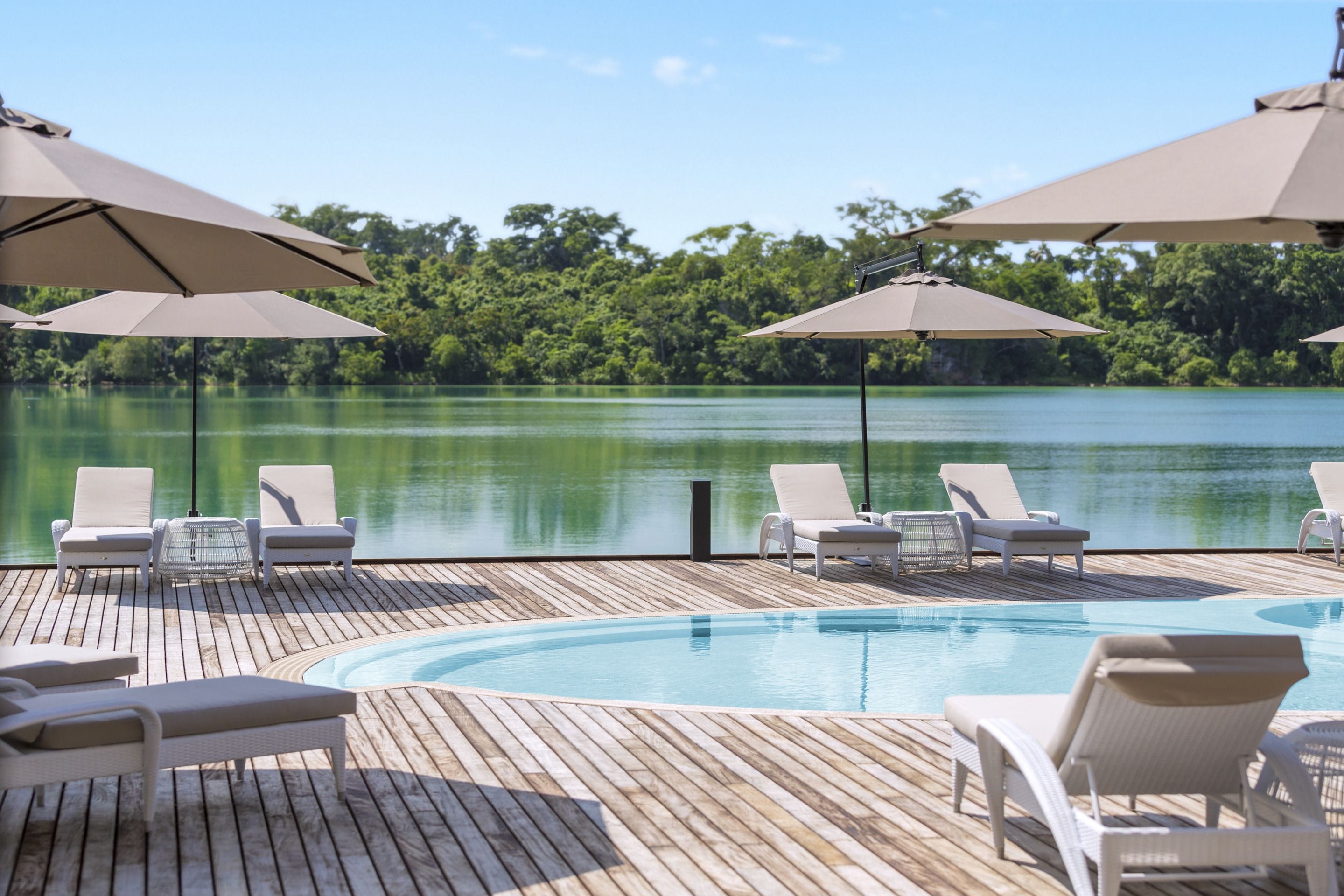 Terraza de madera con piscina, sillas de descanso y sombrillas, con vista a un lago.