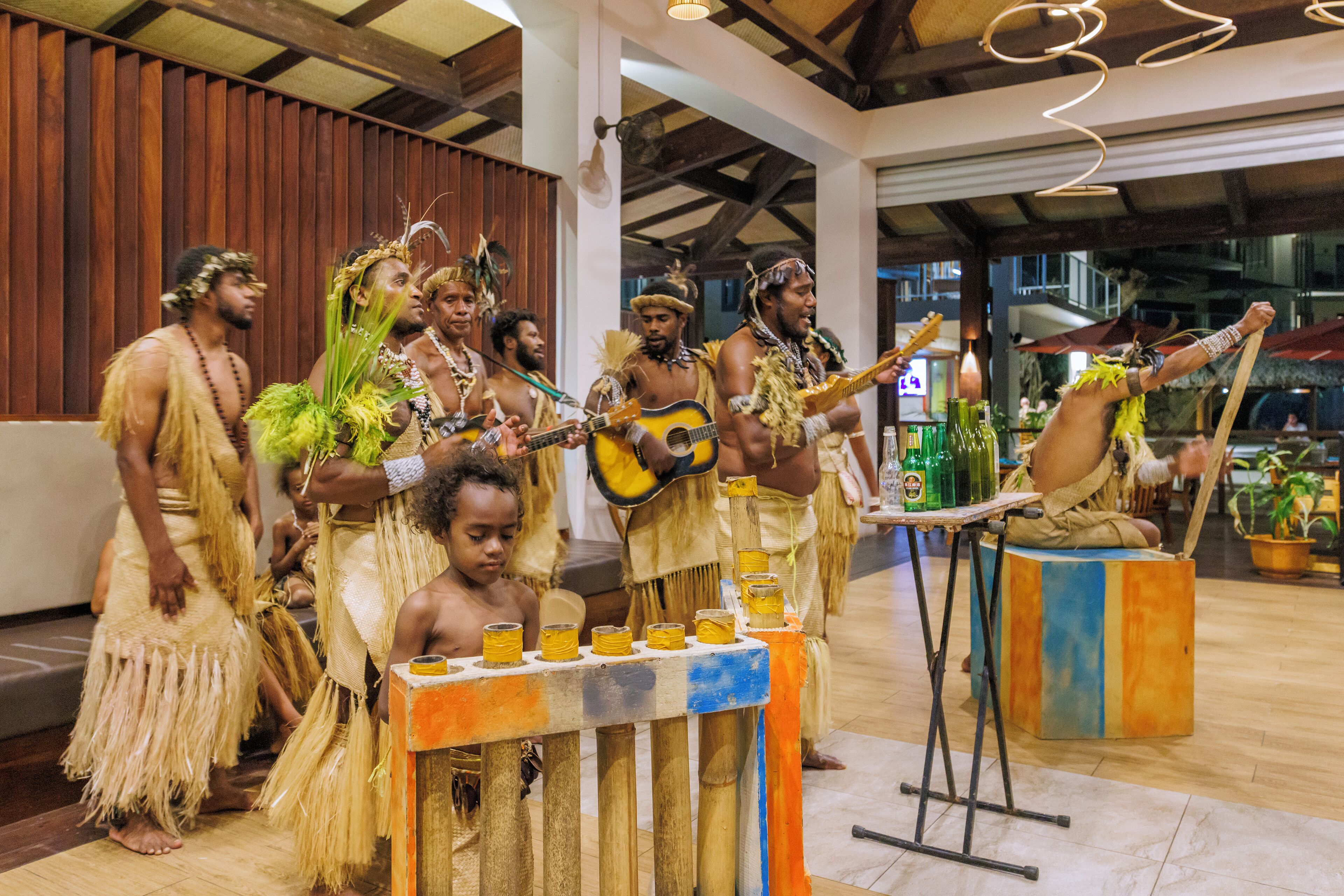 A group of people in traditional attire performing music in a spacious indoor venue with wooden floors and a high ceiling.