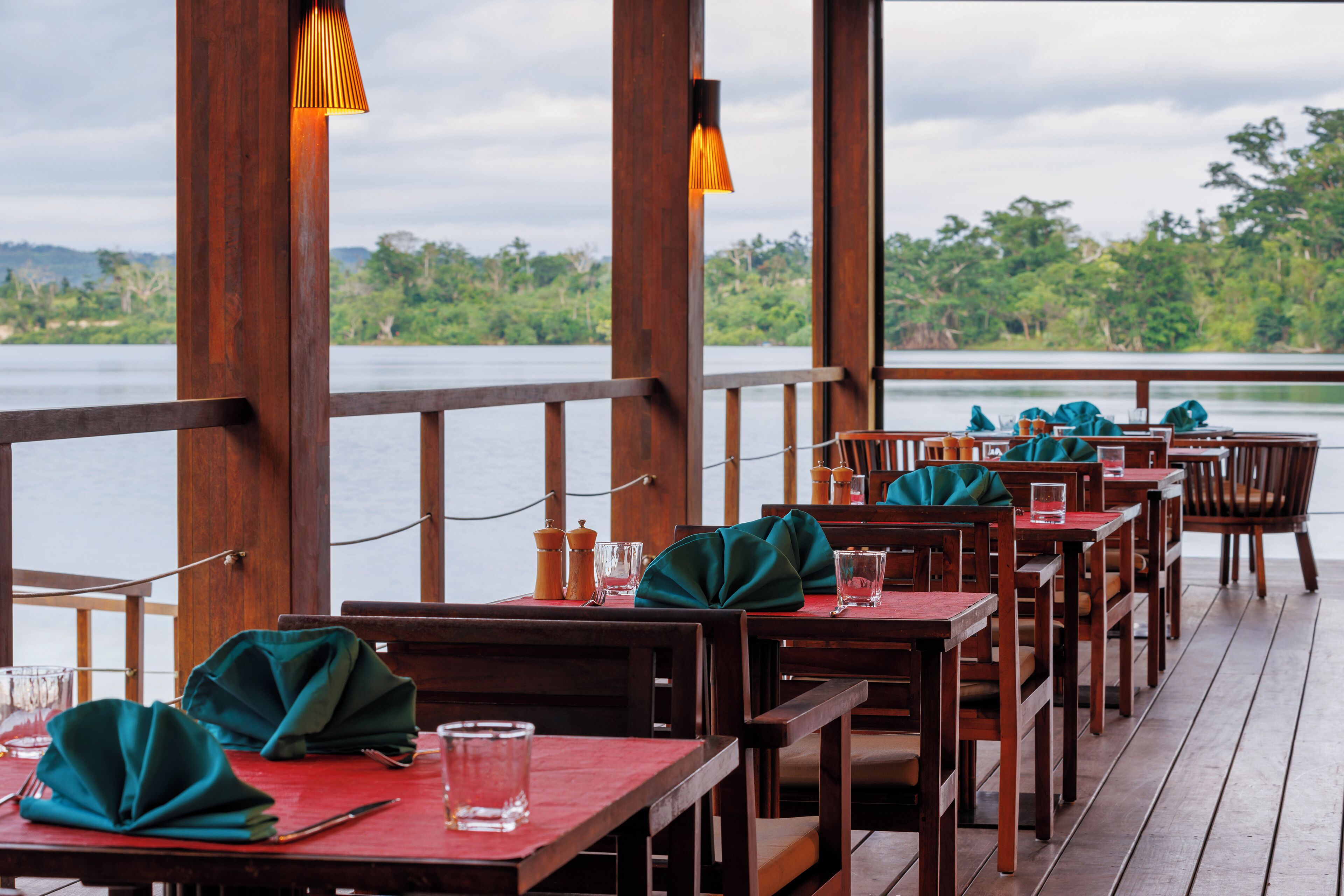 Un restaurant avec des tables et des chaises en bois, soigneusement dressées avec des serviettes vertes et des verres, donnant sur un plan d’eau.